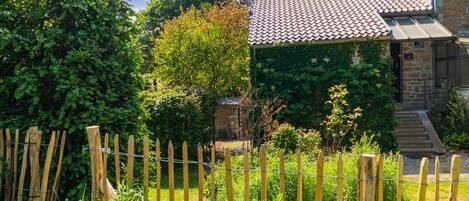 A stone house with a tiled roof, surrounded by lush greenery and a wooden fence.