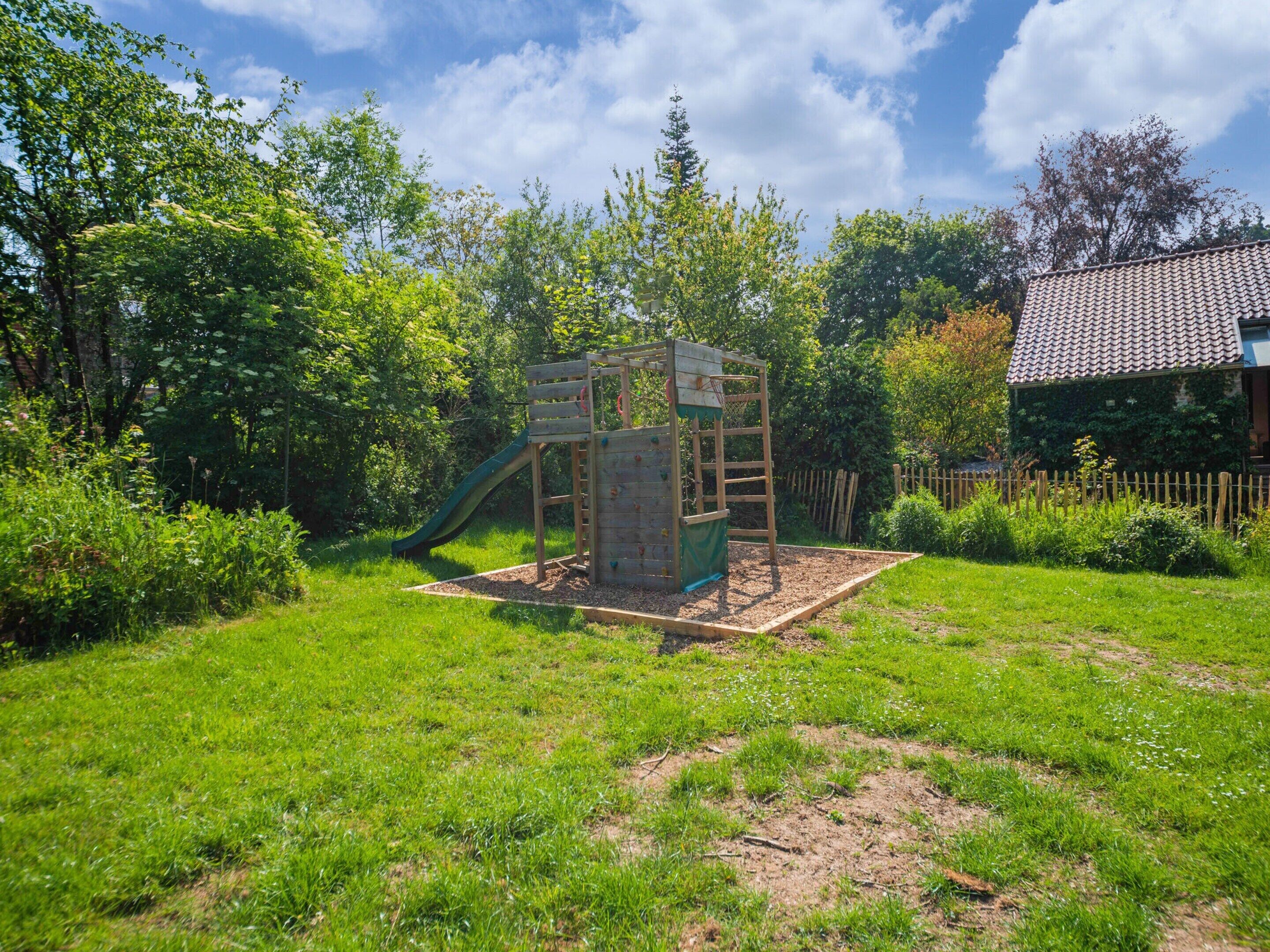 A wooden play structure with a slide and climbing wall in a grassy backyard with a house in the background.