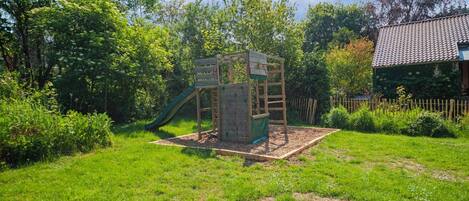 A wooden play structure with a slide and climbing wall in a grassy backyard with a house in the background.