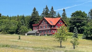 A large, red, multi-story building with a sloped roof, surrounded by a grassy field and dense forest. - Place to Be Modern Retreat (Georgenthal)