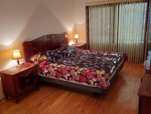 A bedroom with a wooden bed, two bedside tables with lamps, a window with blinds, and a wooden floor.