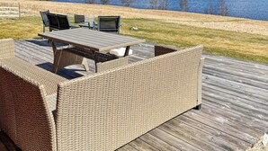 Un área de estar al aire libre con muebles de mimbre, una mesa y sillas, con vistas a un lago y colinas boscosas.
