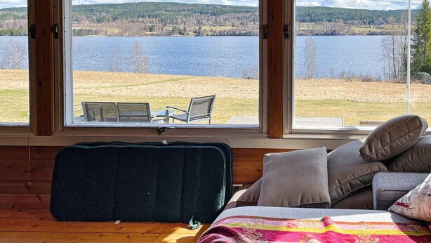 Una habitación con una cama, almohadas y una maleta. Se ven dos ventanas con vistas a un lago y árboles.
