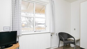 Una habitación con un ventanal grande, un televisor sobre una estantería de madera, una silla de mimbre y un radiador blanco.