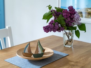 A wooden table with a blue placemat, a glass vase with purple flowers, and two wooden boats on a round wooden tray. - Beach House Dune 19 - Happiness (Ostseebad Dierhagen)
