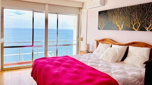 A bedroom with a bed, a nightstand, a painting, and a view of the sea through a sliding door.