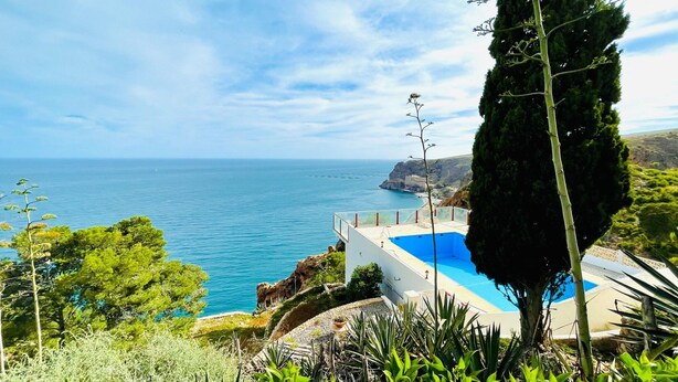 A coastal view with a swimming pool, lush greenery, and a clear sky.