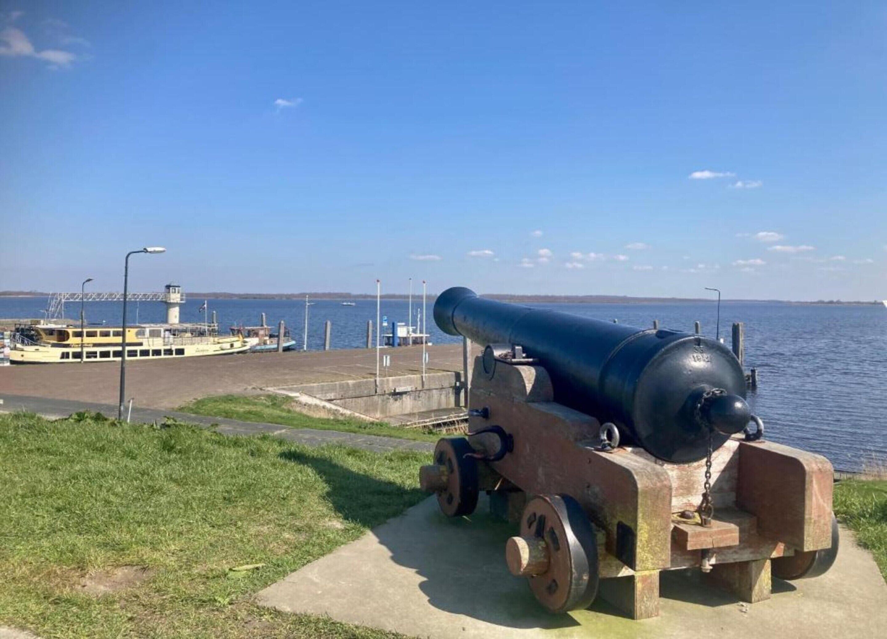 A cannon on a concrete platform by a waterfront with a ferry dock in the background.