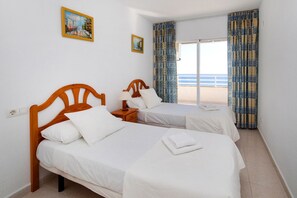 A hotel room with two beds, a nightstand, a lamp, and a view of the sea through a large window.