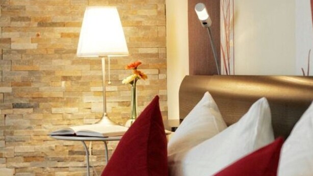 Habitación de hotel con pared de piedra, una mesita de noche con lámpara, un jarrón con flores y almohadas rojas y blancas en la cama.