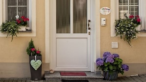 Fachada de edificio con una puerta blanca, dos ventanas con jardineras y una decoración con forma de corazón en el suelo.