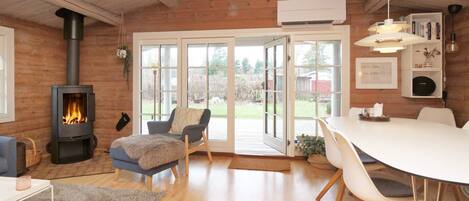 A cozy room with a wood-burning stove, a dining area with a white table and chairs, and a large window with a view of the outdoors.