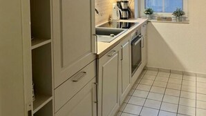 Une cuisine de taille réduite, avec des meubles blancs, un évier, une cuisinière et une fenêtre donnant sur un quartier résidentiel.
