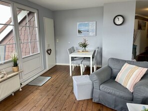 A cozy living room with a grey sofa, a white dining table with chairs, a small white table lamp, and a clock on the wall. - Fehmarn Room With Lake View (Fehmarn)