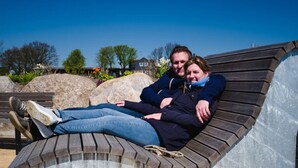 Un couple est assis sur un banc en bois incurvé, avec un mur de pierre en arrière-plan.