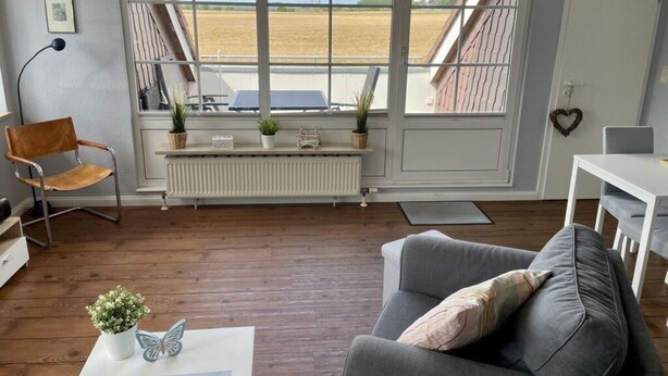 Un salon moderne avec un canapé gris, un plancher en bois, un radiateur et une vue sur un champ, visible à travers de grandes fenêtres.