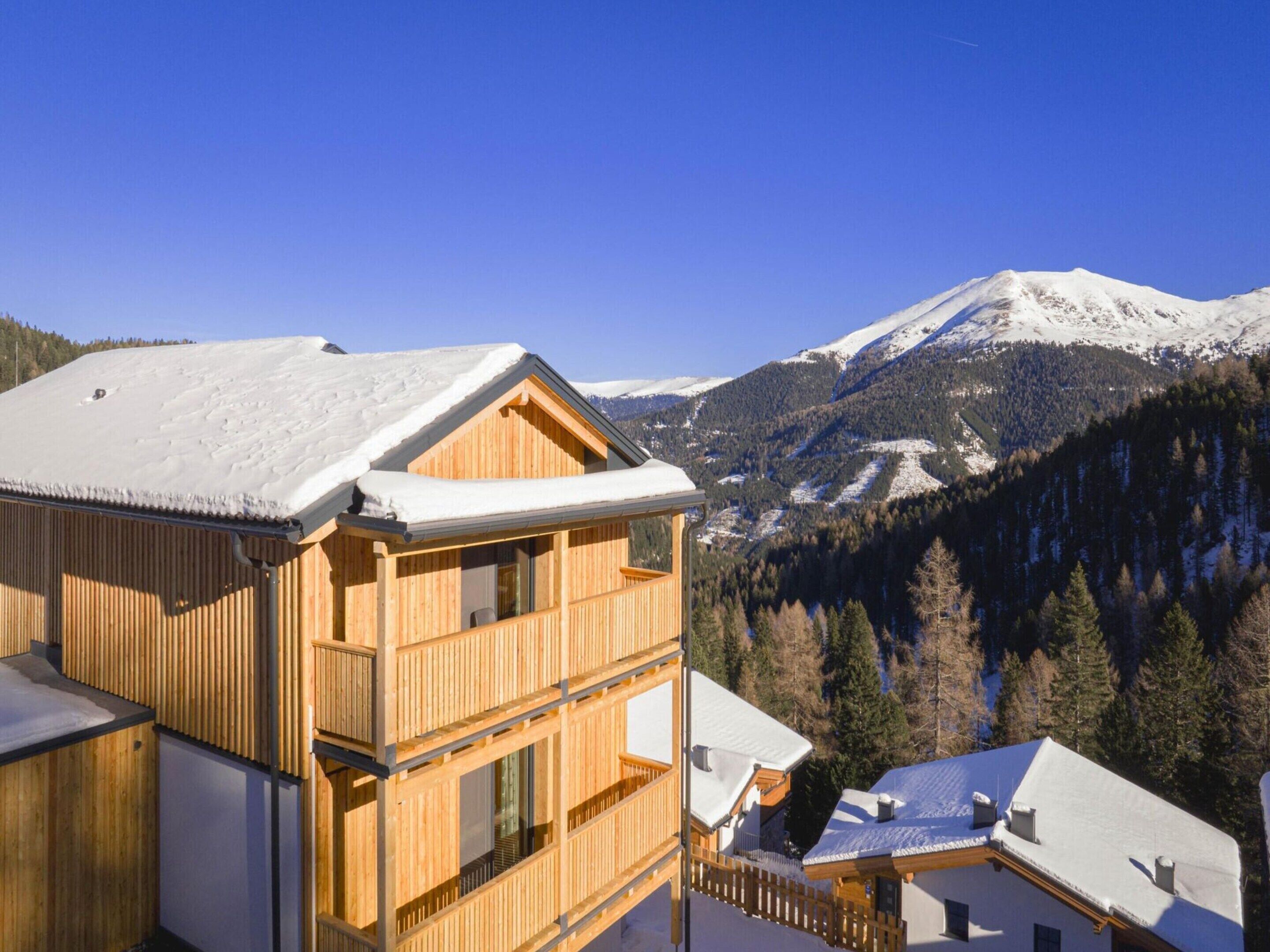 Eine Holzhütte mit Balkon, vor einer verschneiten Berglandschaft und blauem Himmel.