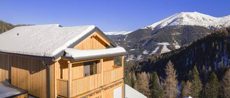 Eine Holzhütte mit Balkon, vor einer verschneiten Berglandschaft und blauem Himmel.