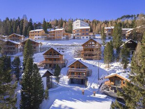 Un albergue cubierto de nieve con varias cabañas de madera, una pista de esquí al fondo y una zona boscosa.