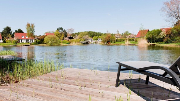 A wooden deck with a bench overlooking a calm lake, surrounded by houses and greenery. - Holiday Home in Groningen With Pool (Vlagtwedde)