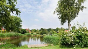 A serene riverside setting with a wooden dock extending into a calm river, surrounded by lush greenery and trees. - Holiday Home in Groningen With Pool (Vlagtwedde)