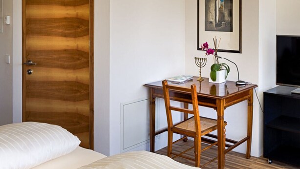Ein Hotelzimmer mit Holztür, einem Bett mit weißer Bettwäsche, einem Holztisch mit Stuhl, einem Fernseher und einem gerahmten Bild an der Wand.