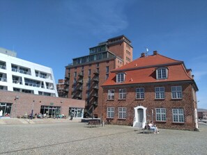 A public square with a cobblestone ground, a red-roofed building with a white entrance, and a modern white building in the background. - Nice Holiday Home With Port View (Wismar)
