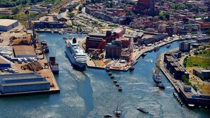 Um porto com um grande navio de cruzeiro atracado, cercado por barcos menores e edifícios industriais.