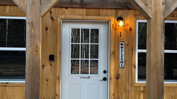 Une cabane en bois avec une porte blanche, un panneau indiquant qu’il est interdit de fumer et un panneau indiquant « Bienvenue ».