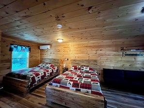 Una cabaña con dos camas de madera, una ventana con persianas, un calentador montado en la pared y un estante con objetos.