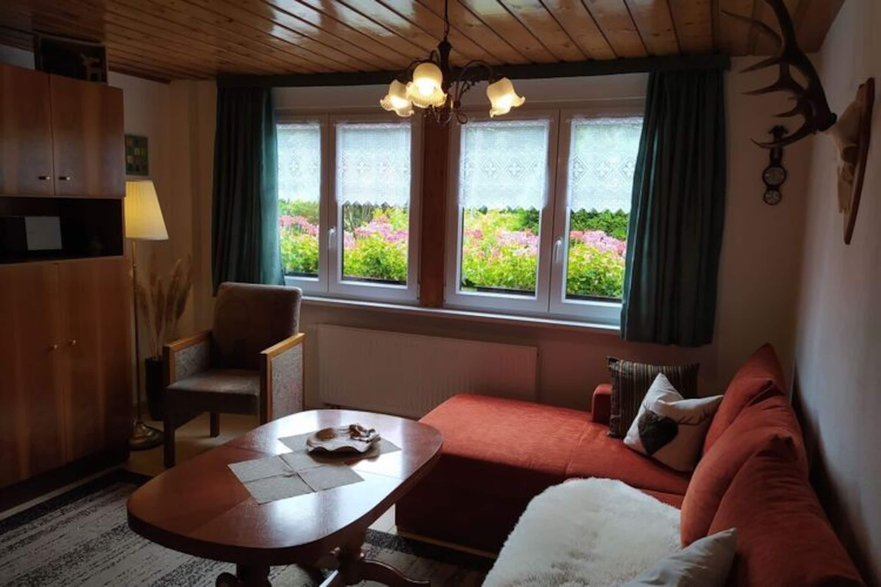 Ein gemütliches Wohnzimmer mit Holzbalkendecke, einem roten Sofa, einer weißen, flauschigen Decke, einem Holztisch und einem Fenster mit Blick auf Grünflächen.