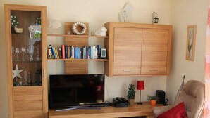 A living room with a TV, wooden cabinets, a bookshelf, a red lamp, and a chair. - Wattloper Right Comfortable Holiday Residence (Wittmund)