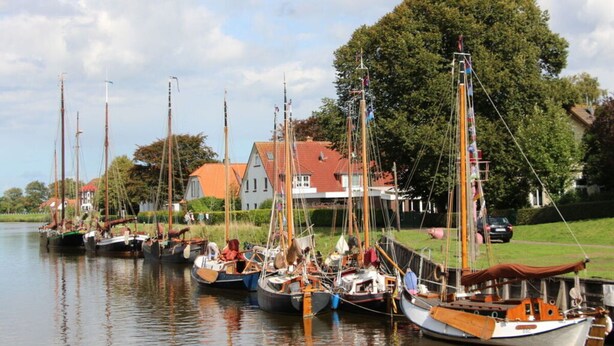 A row of traditional sailing boats docked along a calm river, with houses and trees in the background. - Wattloper Right Comfortable Holiday Residence (Wittmund)