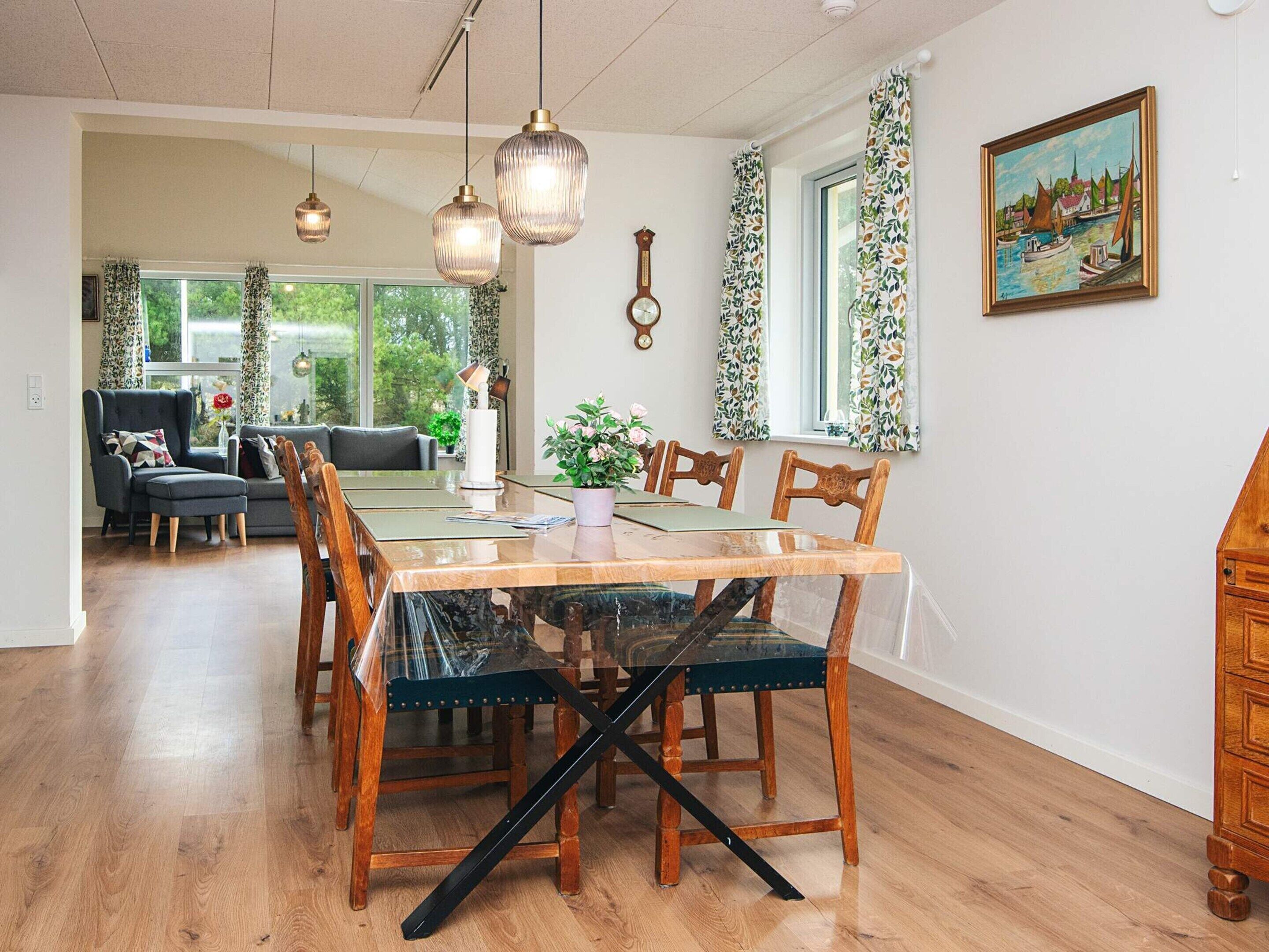 A dining area with a wooden table, chairs, and a glass-top table. There are curtains on the windows, a clock on the wall, and a painting above the table.