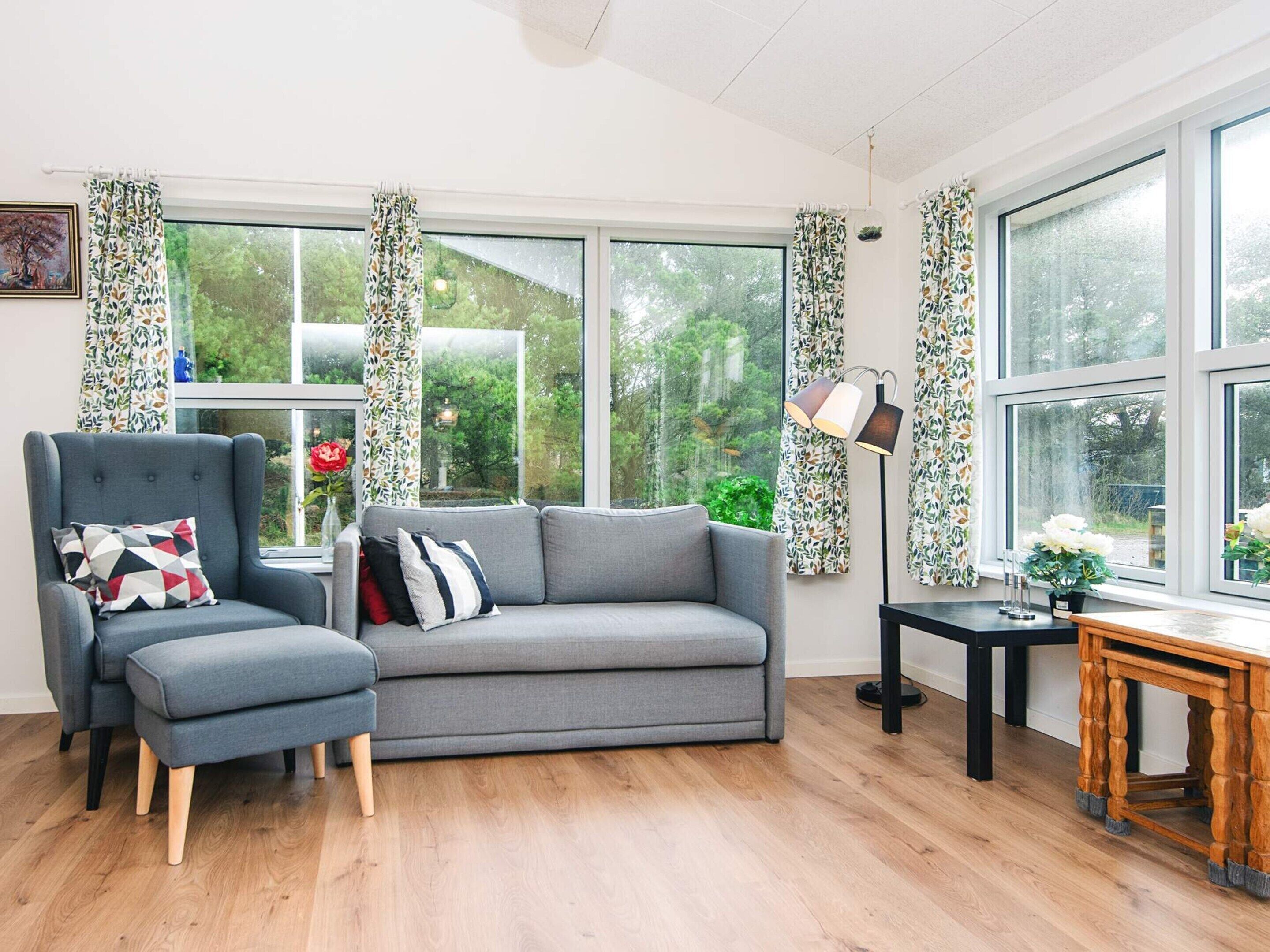 A cozy living room with a grey sofa, matching armchair, and ottoman, a wooden coffee table, and a floor lamp. Large windows with patterned curtains offer a view of greenery outside.