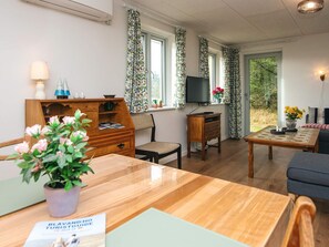 A cozy living room with a wooden table, a sofa, and a TV. There are windows with curtains, a lamp, and a small cabinet. - 8 Person Holiday Home in Blavand (Blavand)