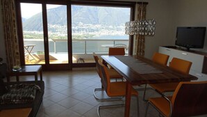 A dining area with a table and chairs, a television, and a view of a lake and mountains through large sliding doors. - Holiday Apartment Casa Chimp 4 (Gambarogno)