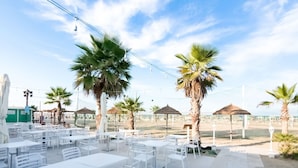 An outdoor patio with white tables and chairs, palm trees, and a view of the beach. - Residence With Swimming Pool Nearby the sea (Rimini)
