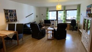 A spacious living area with a wooden floor, a dining table, black sofas, a TV, and a cabinet. - Yvonne Comfortable Holiday Residence (Titisee-Neustadt)