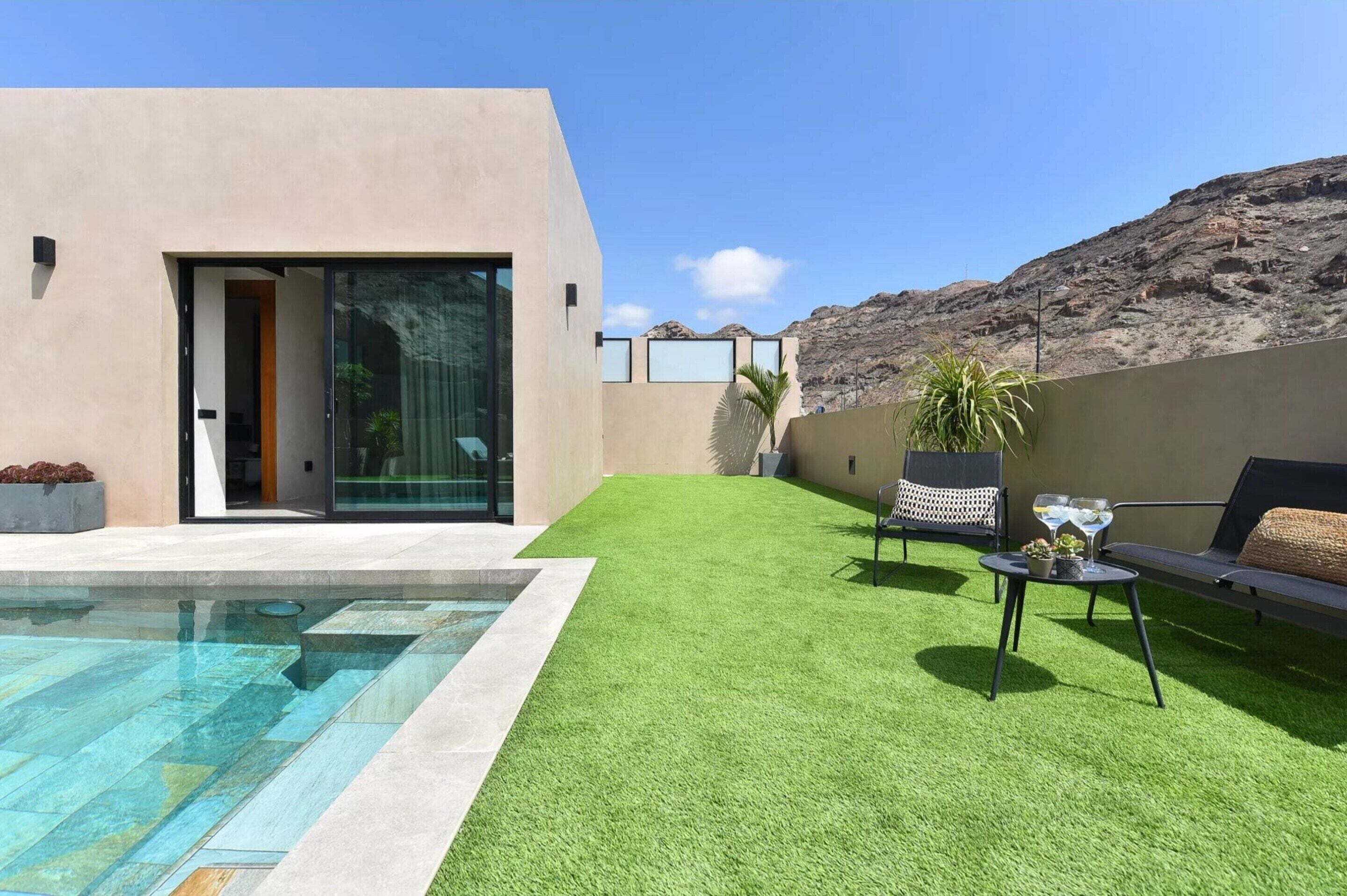 Un espace extérieur moderne doté d’une piscine, d’une pelouse artificielle, d’un coin salon avec un canapé et des fauteuils, et offrant une vue sur un paysage montagneux.
