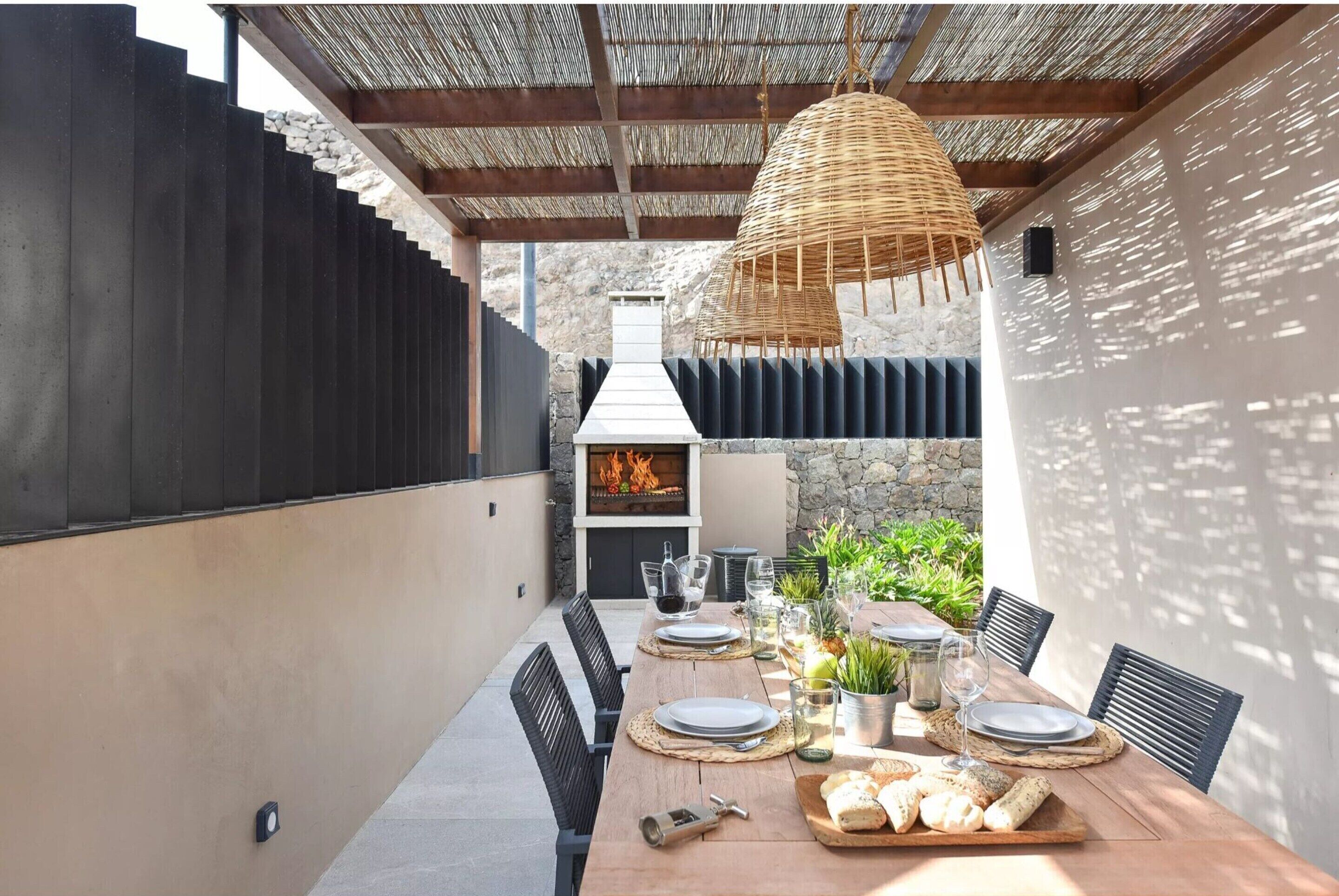 Un espace de restauration en plein air avec une longue table en bois, dressée pour quatre personnes, entourée de chaises noires. Un luminaire tressé est suspendu au-dessus de la table, et un mur de pierre avec un foyer est visible à l’arrière-plan.