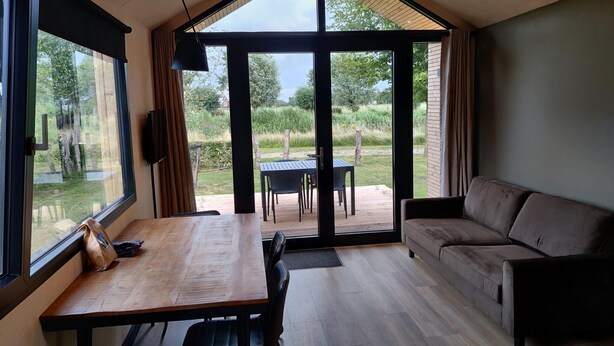 Un interior moderno con una mesa de comedor de madera, sillas negras, un sofá y ventanas amplias con vistas a un jardín.