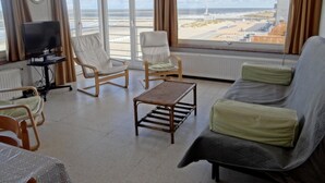 A spacious living room with a large window overlooking a beach and cityscape. There is a sofa, two armchairs, a coffee table, and a TV on a stand. - Apartment in Nieuwpoort With Sea Views (Nieuwpoort)