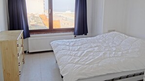A bedroom with a bed, a wooden dresser, a window with curtains, and a radiator. - Apartment in Nieuwpoort With Sea Views (Nieuwpoort)