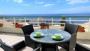 Una mesa redonda con dos platos verdes y copas de vino, rodeada de sillas de mimbre, con vistas a la costa.