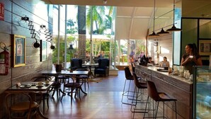 Un restaurante con zona de bar, mesas y sillas de madera. Amplias ventanas dejan entrar luz natural y ofrecen vistas a palmeras.