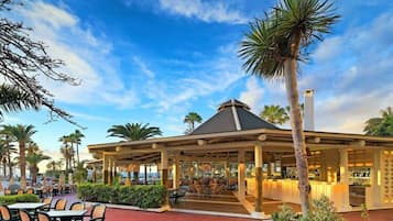 Un restaurante al aire libre con zona de estar techada, palmeras y vista al océano.