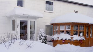 A snow-covered house with a wooden structure in front, possibly a garage or shed, and a small snow-covered bush. - "ganseliesel" Comfortable Holiday Residence (Willingen)