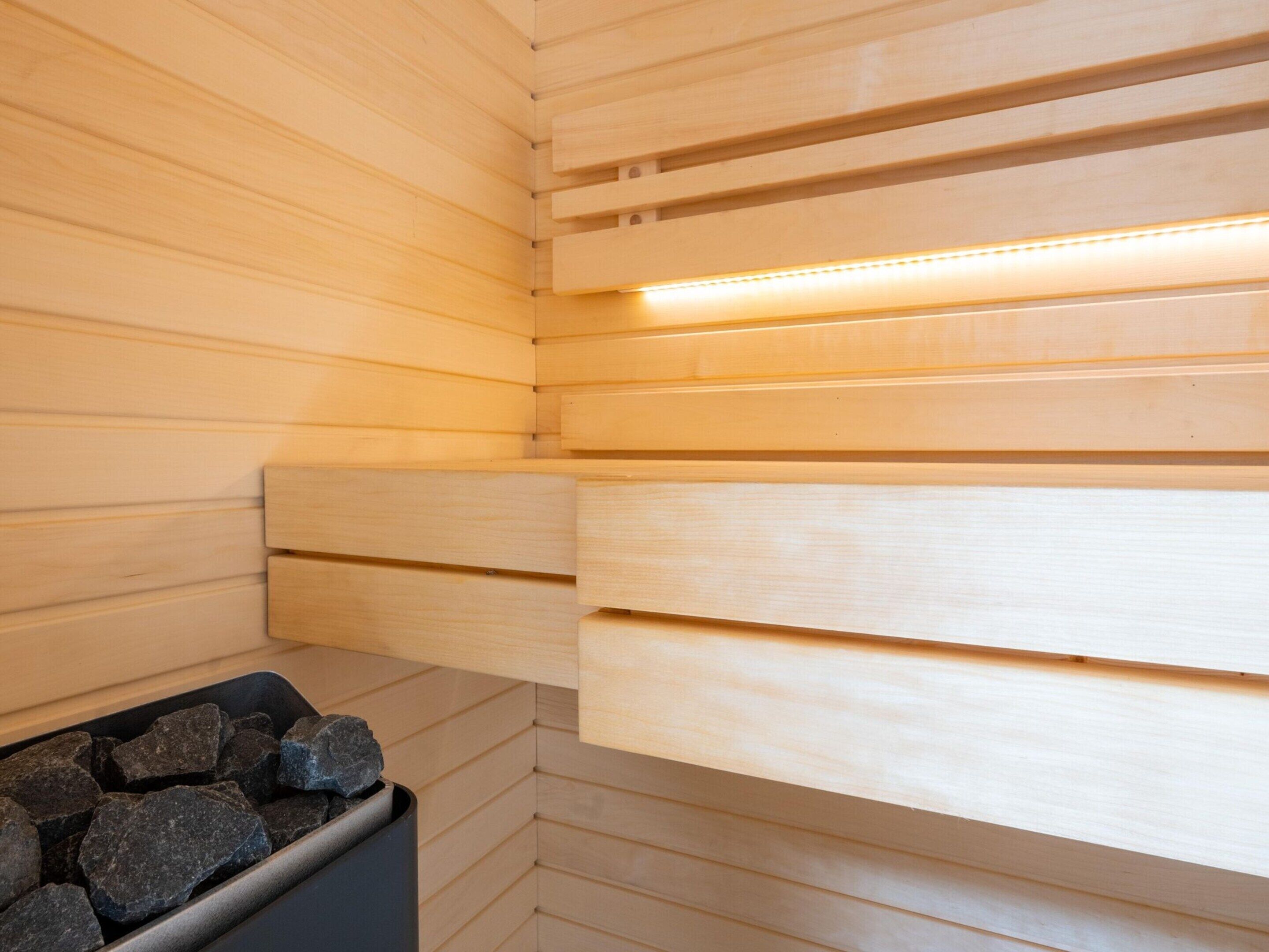 Una sauna con paredes y bancos de madera, un recipiente negro con piedras y una tira de luz a lo largo del techo.