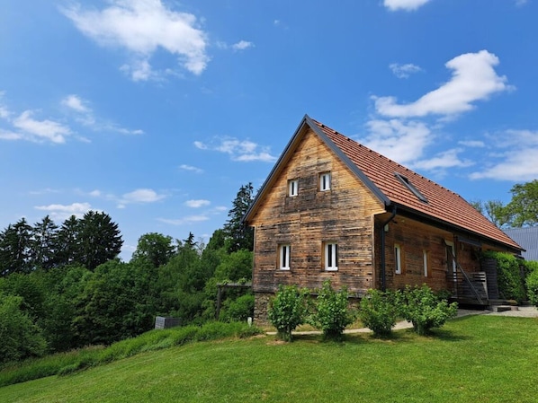 Uma casa de madeira de um andar com telhado inclinado, rodeada por um gramado bem cuidado e árvores densas.